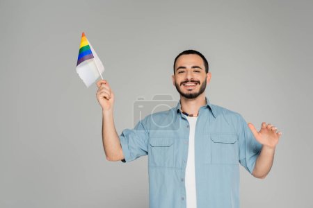 Bearded gay man in shirt smiling and holding rainbow lgbt flag isolated on grey  