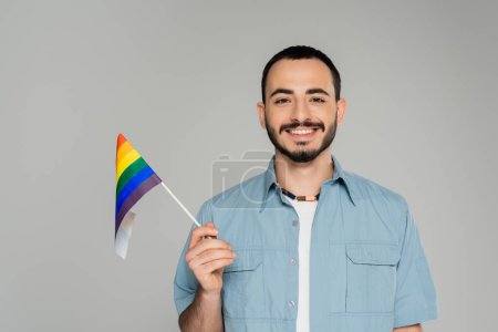 Portrait of smiling and bearded gay man in shirt holding lgbt flag isolated on grey, International Day Against Homophobia 