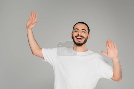 Cheerful gay man in white t-shirt waving hands at camera isolated on grey  