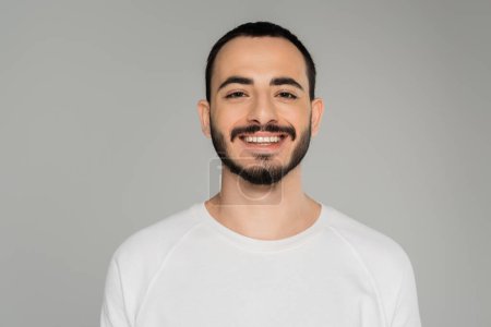 Porträt eines lächelnden schwulen Mannes im weißen T-Shirt, der isoliert auf grau in die Kamera blickt  