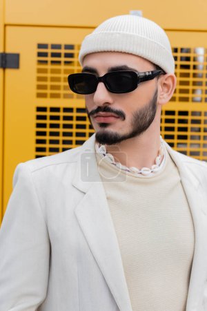 Portrait of fashionable gay man in hat and sunglasses standing on urban street 