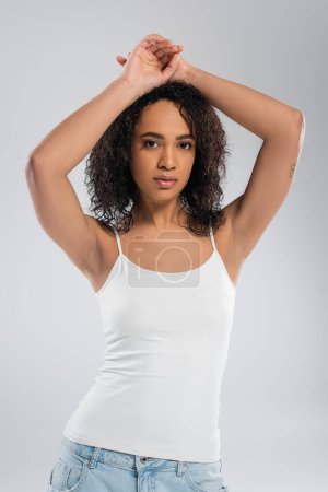 brunette african american woman in white tank top posing with hands above head isolated on grey