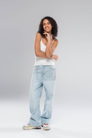 Photo for Full length of joyful african american woman in white tank top and blue jeans on grey background - Royalty Free Image