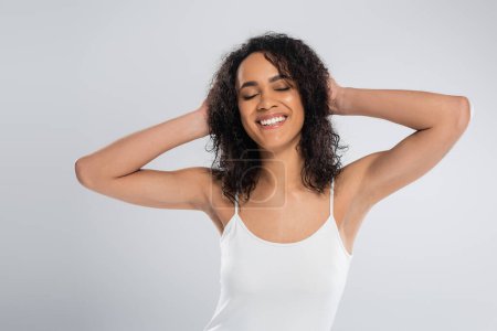 carefree african american woman in white tank top touching wavy hair and smiling with closed eyes isolated on grey