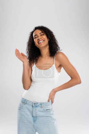 joyful african american woman in white tank top waving hand and smiling isolated on grey