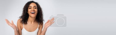 excited african american woman in white tank top showing wow gesture and looking at camera isolated on grey, banner