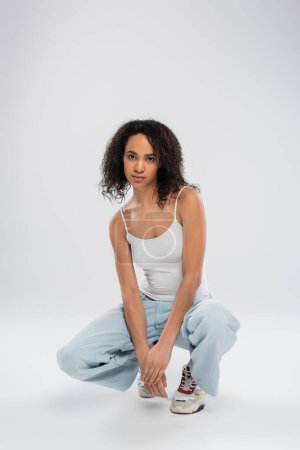 full length of african american woman in white tank top and jeans with sneakers sitting on haunches on grey background