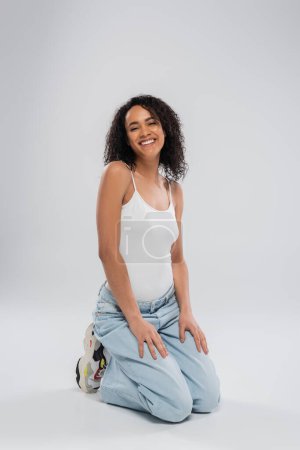 full length of pretty african american woman in jeans and white tank top sitting on haunches on grey background