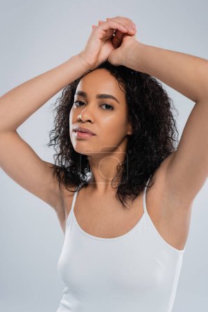 Téléchargez les photos : Portrait de jeune femme afro-américaine aux cheveux bruns bouclés posant les mains au-dessus de la tête isolée sur gris - en image libre de droit
