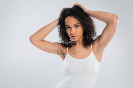 young african american woman in white tank top touching wavy brunette hair and looking at camera isolated on grey