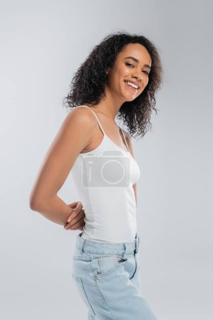 brunette african american woman in white tank top posing with hands behind back and smiling at camera isolated on grey