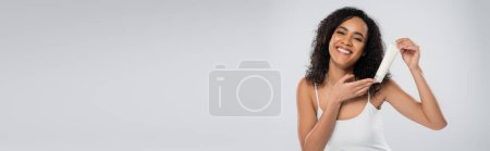 positive african american woman showing tube with cosmetic cream isolated on grey, banner