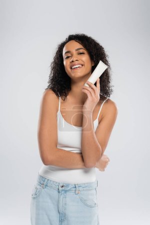 pleased african american woman in white tank top posing with tube of cosmetic cream isolated on grey