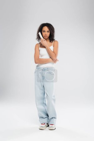 full length of african american woman in white tank top and blue jeans holding cosmetic cream on grey background