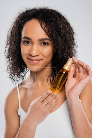 pleased african american woman showing hair oil and smiling at camera isolated on grey