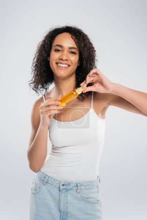 excited african american woman holding bottle of cosmetic oil while looking at camera isolated on grey