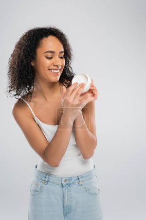mujer afroamericana complacida en camiseta sin mangas mirando contenedor de crema cosmética aislado en gris