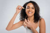 joyful african american woman applying cosmetic serum on perfect face isolated on grey mug #652414562
