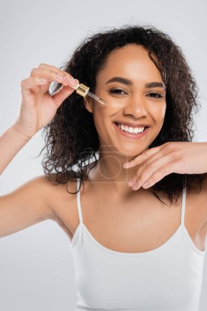 pleased african american woman looking at camera and applying cosmetic serum isolated on grey