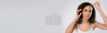brunette african american woman in white tank top applying cosmetic serum isolated on grey, banner