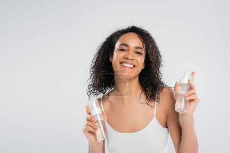 young african american woman posing with bottles of face foam while smiling at camera isolated on grey