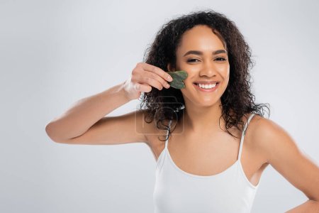 happy african american woman doing face massage with jade scraper isolated on grey