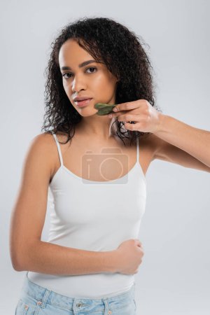 mujer joven afroamericana en camiseta blanca con raspador facial aislado en gris