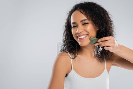 brunette african american woman doing lifting massage with face scraper isolated on grey