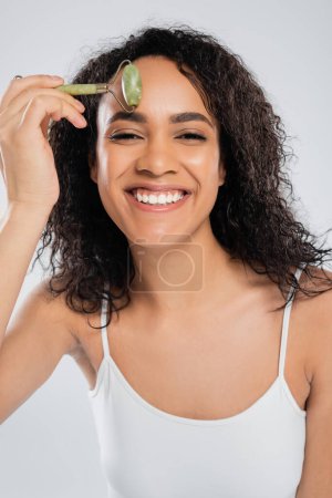 cheerful african american woman doing face massage with jade roller isolated on grey