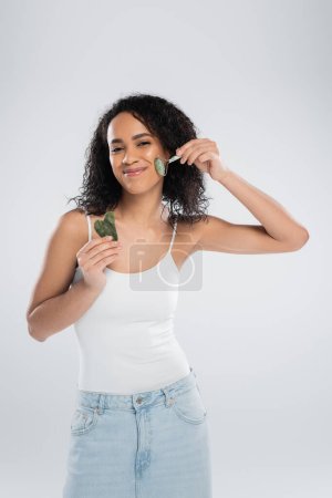pleased african american woman holding face scraper and using jade roller isolated on grey