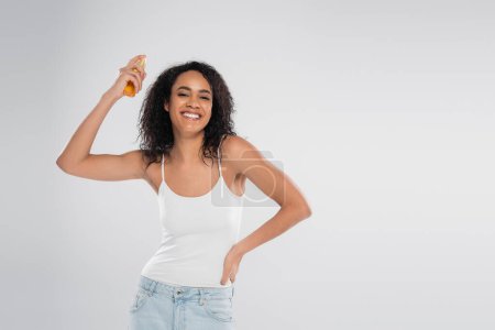 femme afro-américaine souriante appliquant de l'huile capillaire tout en se tenant debout avec la main sur la hanche isolé sur gris