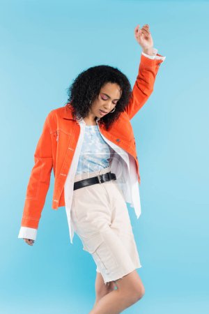 mujer afroamericana de moda en chaqueta naranja brillante posando con la mano levantada aislada en azul