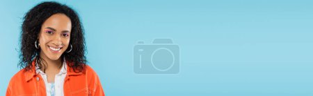 Photo for Portrait of pleased african american woman with wavy brunette hair smiling at camera isolated on blue, banner - Royalty Free Image