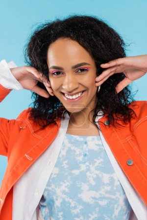 Porträt einer fröhlichen und stilvollen afrikanisch-amerikanischen Frau mit hellem Make-up, die Hände in der Nähe des Gesichts hält, isoliert auf blauem Grund