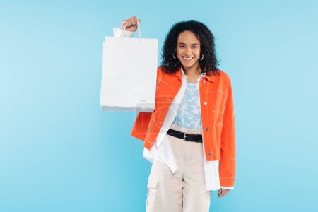 überglückliche Afroamerikanerin in orangefarbener Jacke mit weißen Einkaufstaschen auf blauem Grund