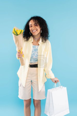 Photo for Pleased african american woman in stylish clothes posing with yellow tulips and shopping bags isolated on blue - Royalty Free Image