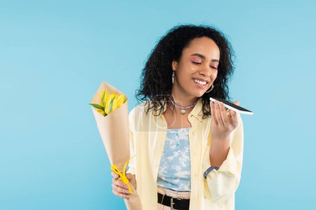 positive african american woman with yellow tulips sending voice message on smartphone isolated on blue
