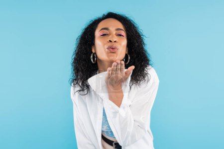 Téléchargez les photos : Brune afro-américaine femme souffler air baiser et regarder caméra isolé sur bleu - en image libre de droit