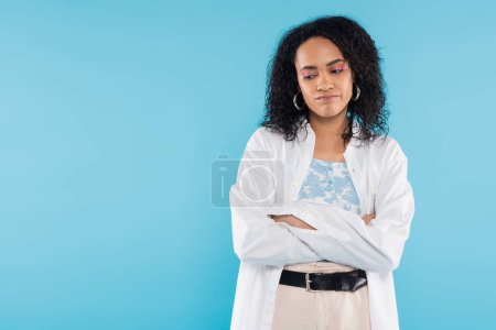upset african american woman in white shirt standing with folded arms isolated on blue