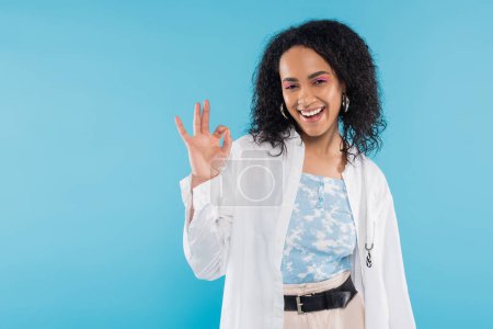 femme afro-américaine ravie en chemise élégante souriant à la caméra et montrant OK signe isolé sur bleu
