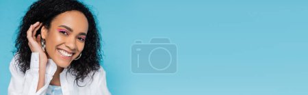 Téléchargez les photos : Portrait de femme afro-américaine heureuse avec une peau parfaite fixant les cheveux ondulés et souriant à la caméra isolée sur bleu, bannière - en image libre de droit