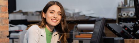 cheerful woman smiling near monitor while working in print center, banner 