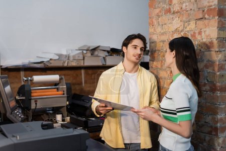 cheerful man and woman holding laminated paper near print plotter in copy center 