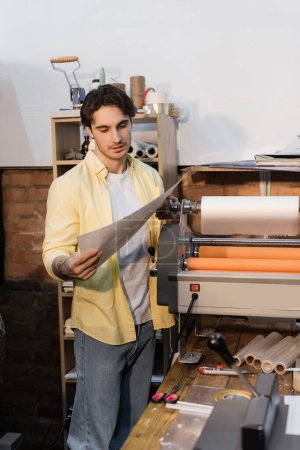 tipógrafo guapo mirando papel impreso cerca de plotter de impresión