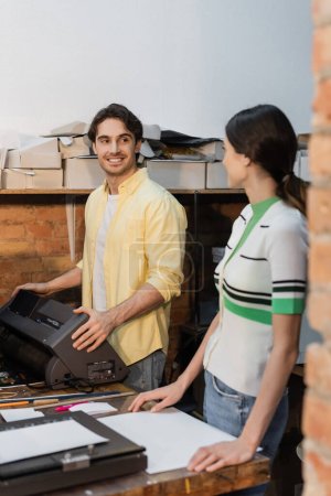 cheerful printing operator looking at colleague while standing near printer 