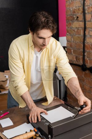 Téléchargez les photos : Homme tatoué utilisant une machine de coupe-papier dans le centre d'impression moderne - en image libre de droit