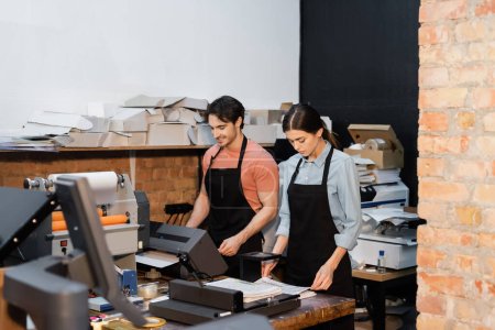 cheerful man in apron working with printer next to pretty colleague in print center 