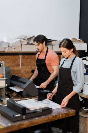 handsome worker in apron using printer next to pretty colleague looking at newspapers in print center 
