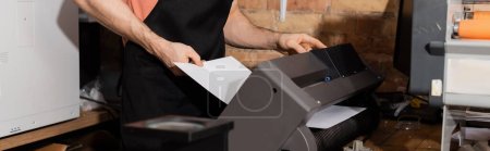 vista recortada del trabajador en delantal sosteniendo papel cerca de la impresora y periódicos frescos en el centro de impresión, pancarta 