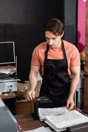 Photo for Tattooed man in apron holding newspaper near magnifying glass in print center - Royalty Free Image